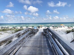 A wooden boardwalk leading to a beach with umbrellas and people. - 🏖️ Emerald Isle 1402 | 14th Floor Beachfront | 2 Pools + Walk to Pier (Gulf Breeze)