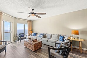 A spacious living room with a large window, a ceiling fan, a sofa with striped cushions, a wooden coffee table, and a side table with a lamp. - 🏖️ Emerald Isle 1402 | 14th Floor Beachfront | 2 Pools + Walk to Pier (Gulf Breeze)