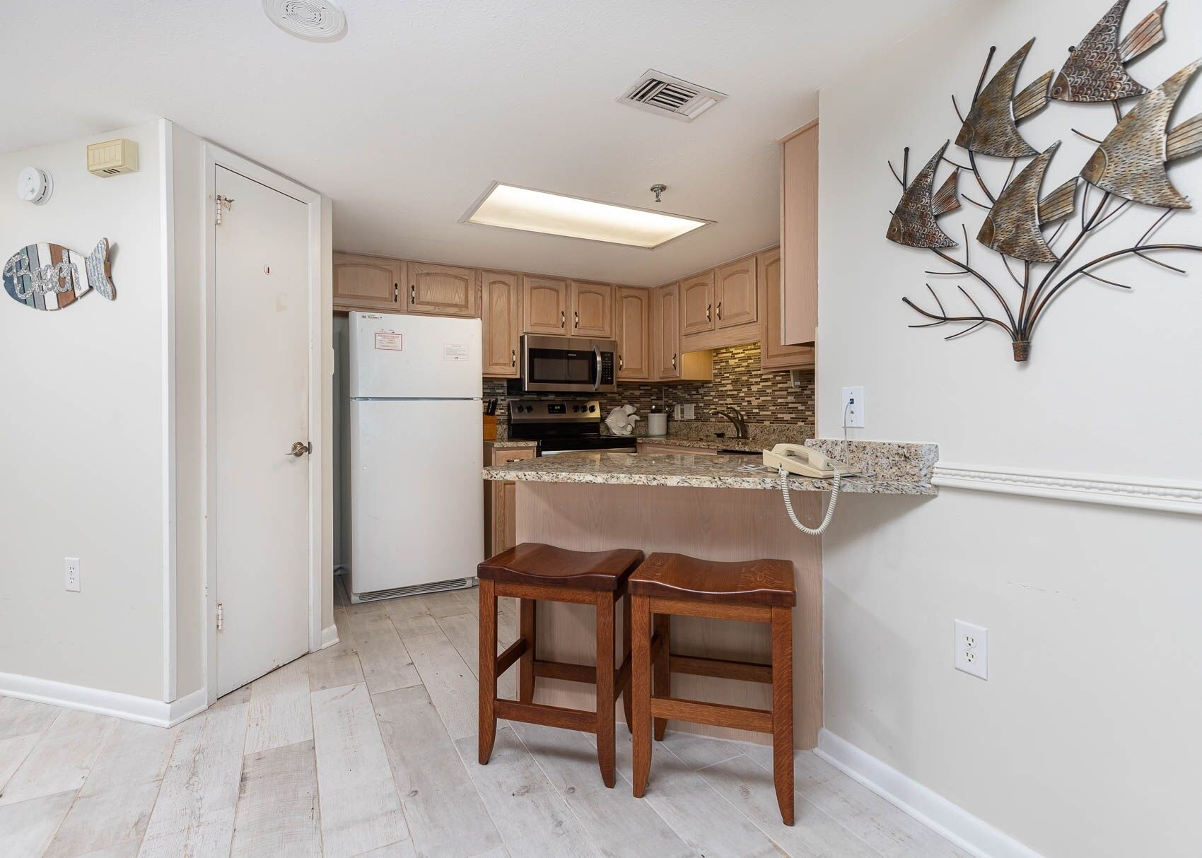 A kitchen with wooden cabinets, a refrigerator, a microwave, and a granite countertop. There are two wooden stools and a decorative metal wall piece.