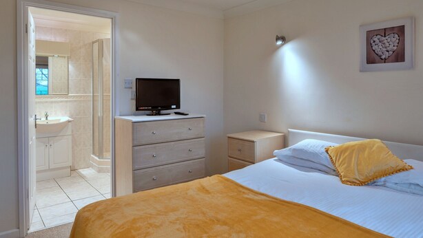 Ein Schlafzimmer mit Bett, Nachttisch, Fernseher auf einer Kommode, Badezimmer mit Waschbecken und Spiegel sowie einer Wand mit Herzdekoration.