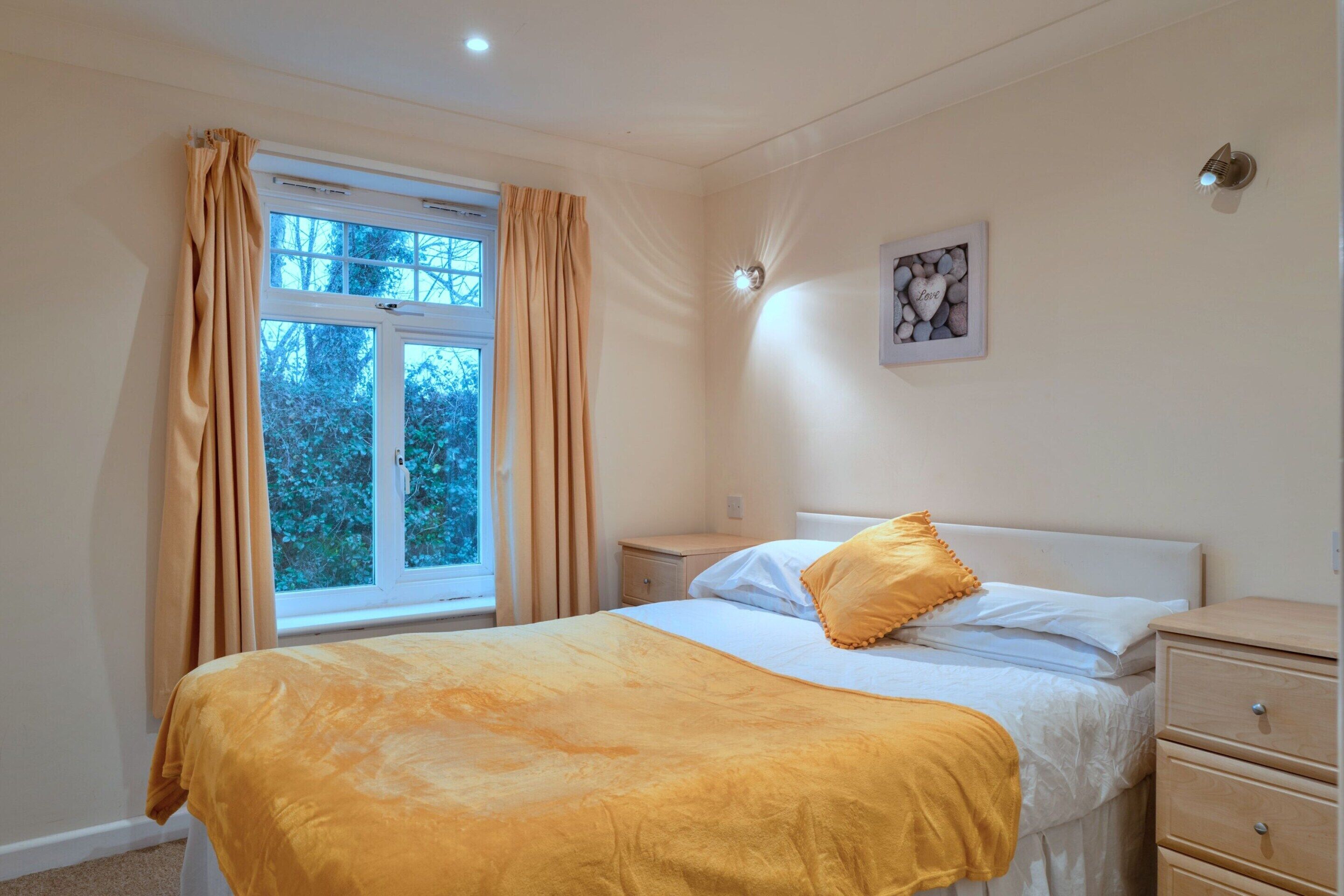 Quarto com cama, mesa de cabeceira, luminária na parede e cortinas na janela.