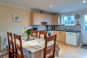 A kitchen with wooden cabinets, a dining table with chairs, a vase of flowers, and a clock on the wall. - Spacious Holiday Home in Newquay (St. Columb Major)