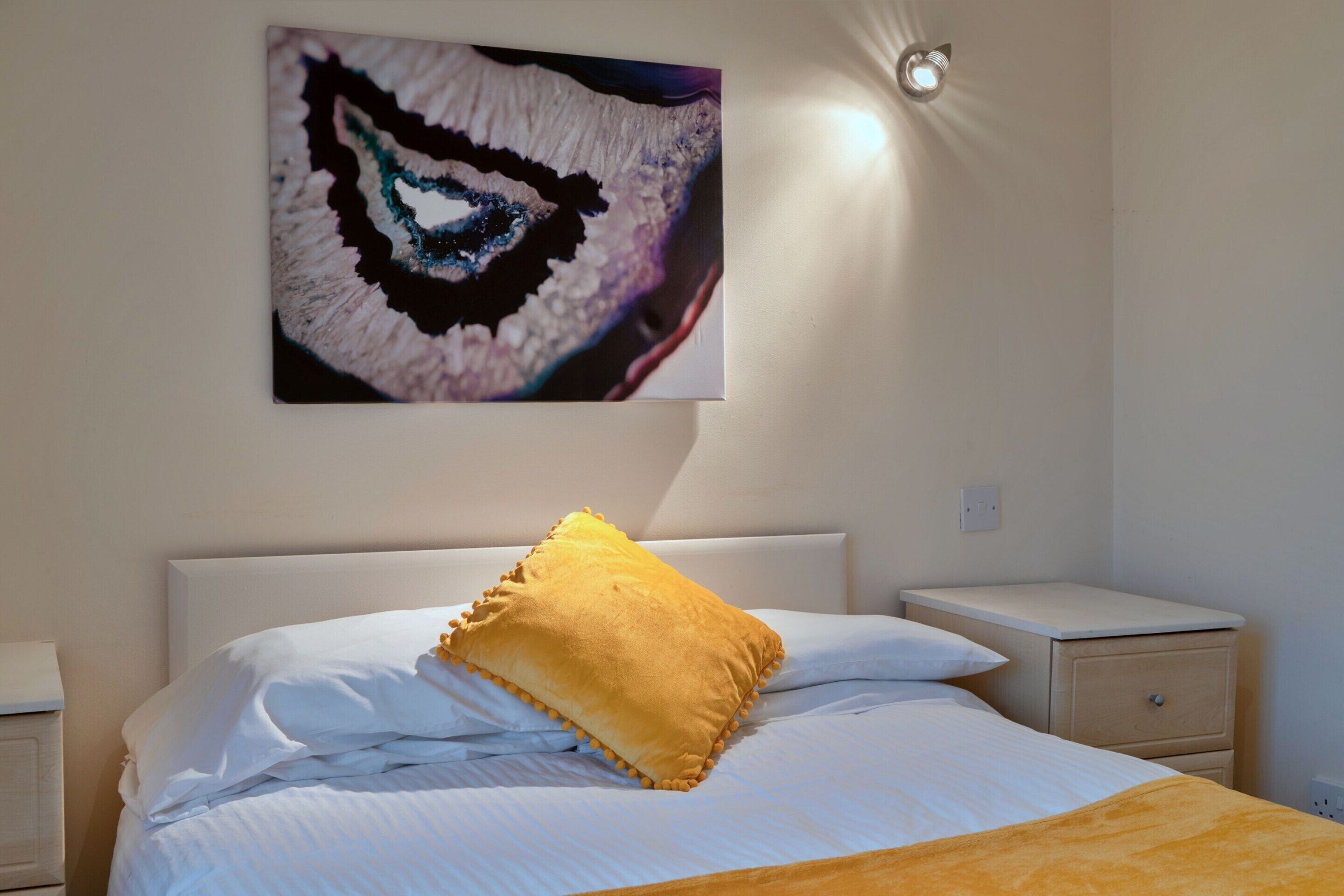 A hotel room with a bed, a nightstand, a wall-mounted light, and a piece of abstract art.