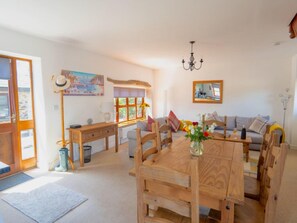 A cozy living room with a wooden dining table, a sofa, and a large window. - 3-bedroom Cottage Near Coast (Bude)
