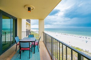 A balcony with a table and chairs, overlooking a beach with people and umbrellas. - Phoenix Iii 3067 1 Bedroom Condo (Orange Beach)