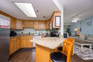 A kitchen with wooden cabinets, granite countertops, a refrigerator, and a dining area with a chair and table. - Phoenix Iii 3095 1 Bedroom Condo (Orange Beach)