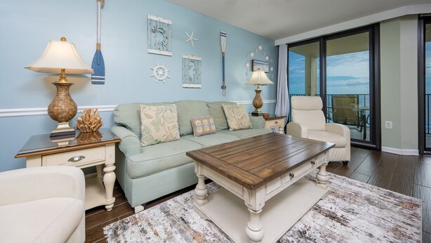 A living room with a light blue wall, a wooden coffee table, a sofa with patterned cushions, a white armchair, and a ceiling fan. - Phoenix Iii 3095 1 Bedroom Condo (Orange Beach)