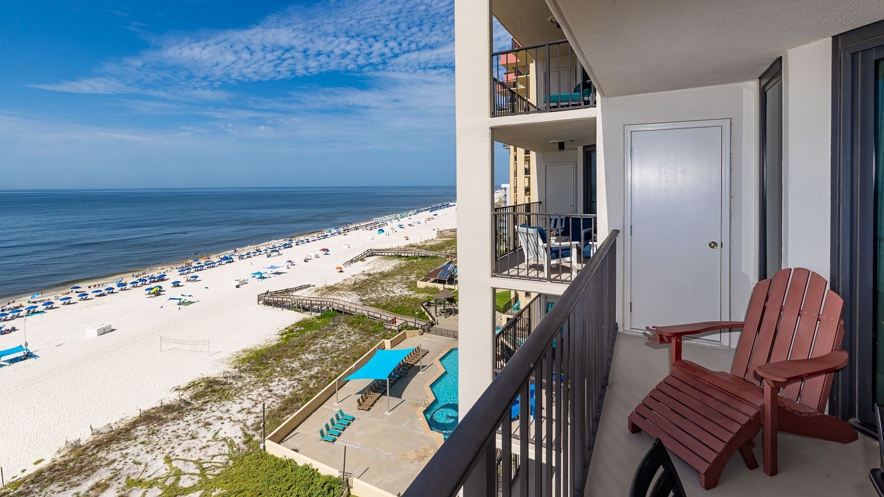 A balcony with a view of a beach, a pool, and a red Adirondack chair.