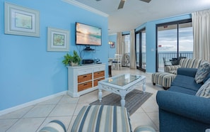 A living room with a television, a white cabinet with baskets, a glass coffee table, striped sofas, and a view of the ocean through large windows. - Phoenix X 505 1 Bedroom Condo (Orange Beach)