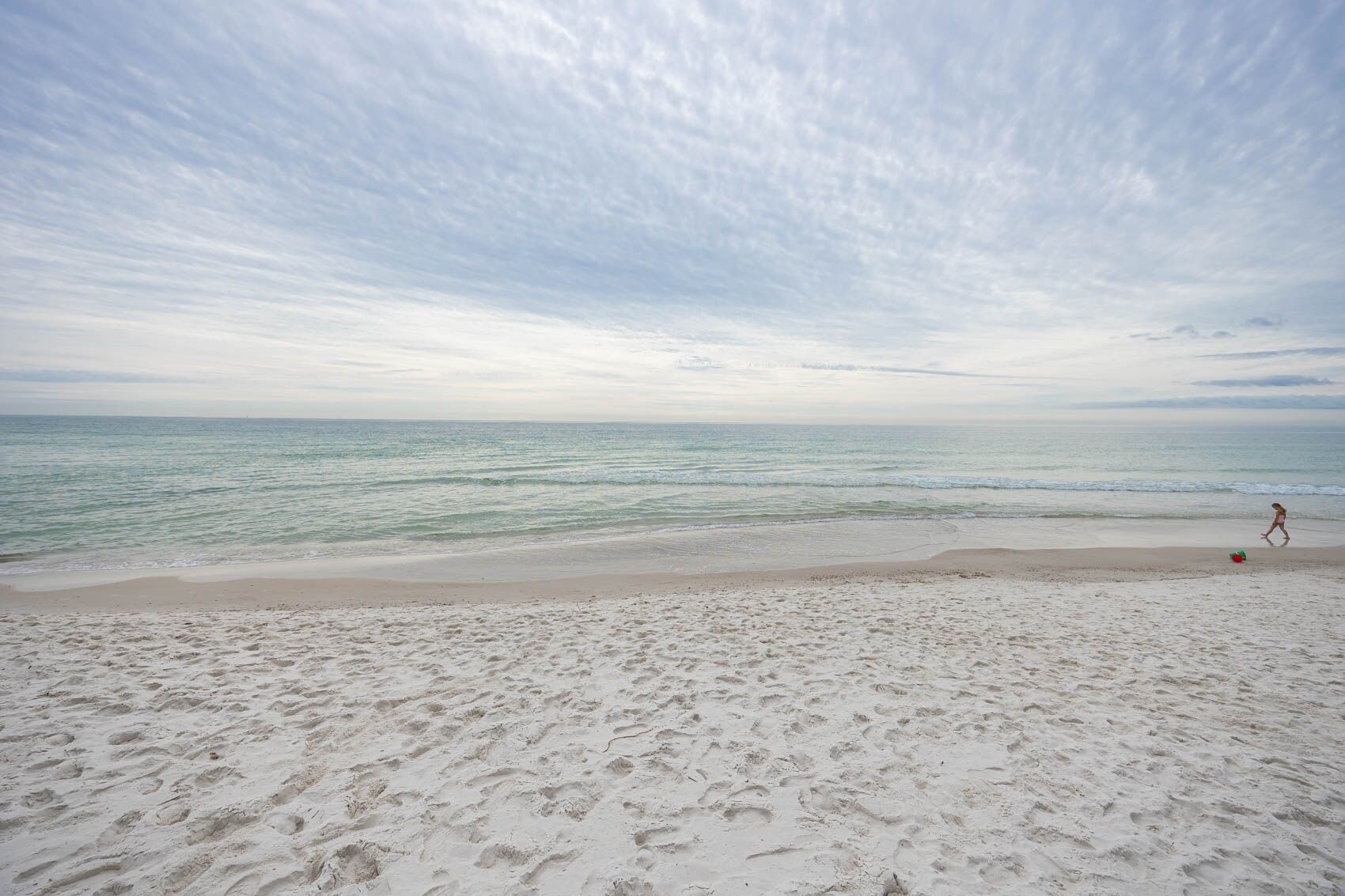 Une plage où l’on voit des empreintes de pas menant à l’eau, un enfant qui joue et un ciel nuageux.