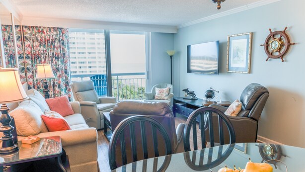 A modern living room with a sofa, armchairs, a coffee table, and a dining table. There is a TV mounted on the wall, a lamp, and a decorative wall clock. - 705 S Hampton Oceanview Retreat With Balcony 2 Bedroom Condo (Myrtle Beach)