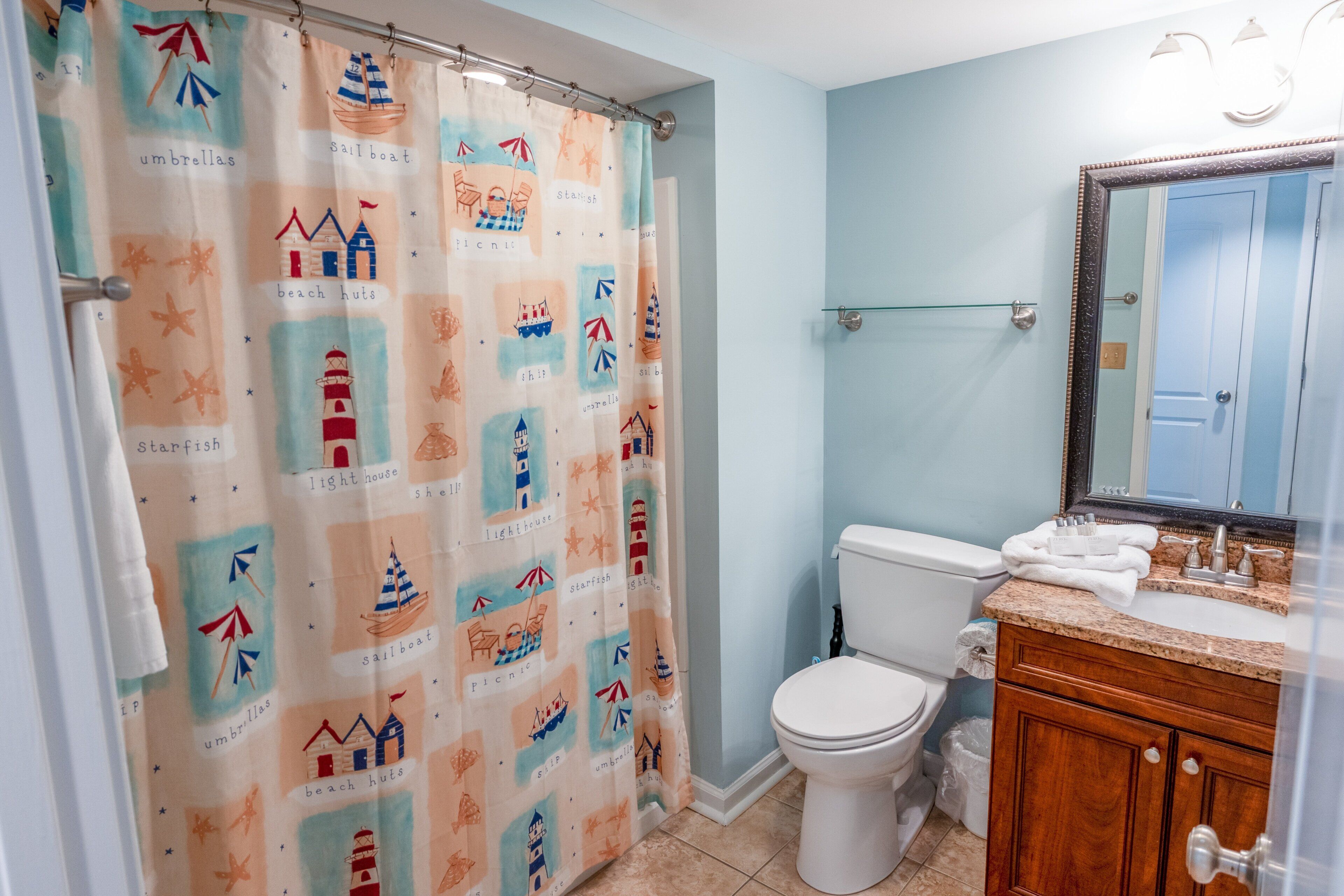 Un baño con cortina de ducha de motivos náuticos, un inodoro, un lavabo con espejo y un lavamanos.