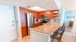 A modern kitchen with a stainless steel refrigerator, a microwave, and a stove. There is a granite countertop and a bar stool area. - 705 S Hampton Oceanview Retreat With Balcony 2 Bedroom Condo (Myrtle Beach)