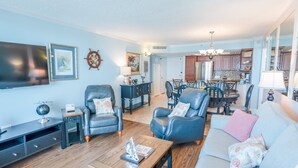 A spacious living room with a sofa, two armchairs, a coffee table, and a television. There is a dining area with a table and chairs in the background. - 705 S Hampton Oceanview Retreat With Balcony 2 Bedroom Condo (Myrtle Beach)