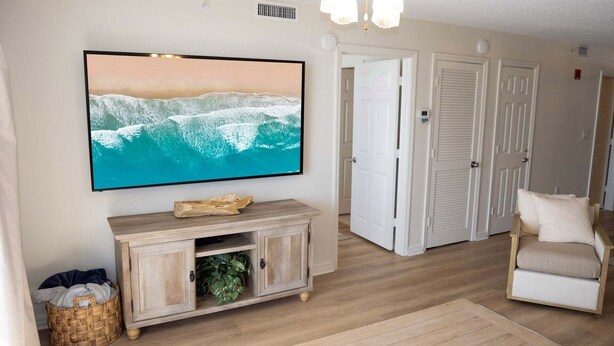A living room with a television mounted on the wall, a wooden cabinet, a wicker basket, and a beige armchair. - Pelican Beach 1013 2 Bedroom Condo (Destin)