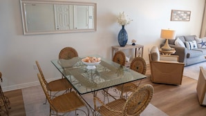 A dining area with a glass table, wicker chairs, a bowl of snacks, a vase with flowers, and a sofa with cushions. - Pelican Beach 1013 2 Bedroom Condo (Destin)