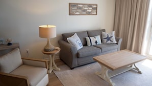 A living room with a sofa, a coffee table, a lamp, and a wall art piece. - Pelican Beach 1013 2 Bedroom Condo (Destin)