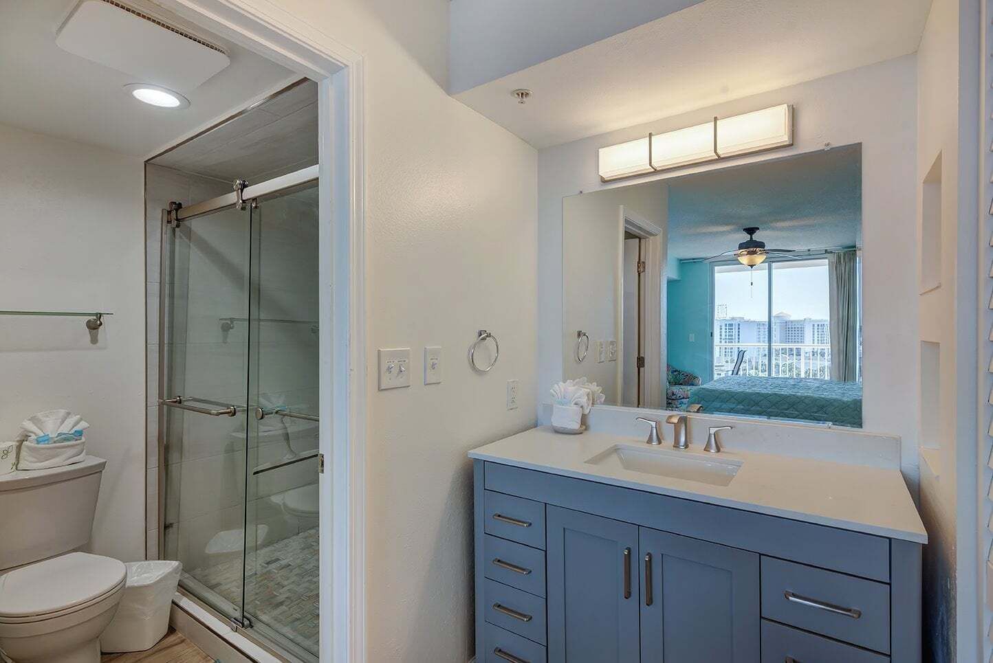 A modern bathroom with a glass shower enclosure, a white toilet, a vanity with a mirror, and a view of a cityscape through the window.