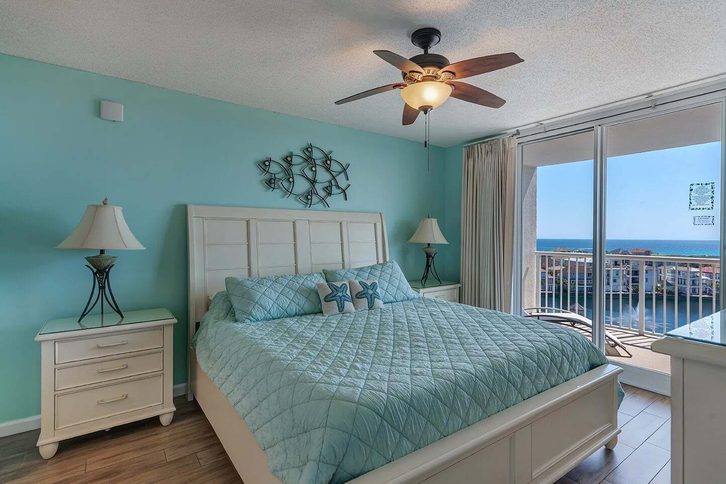Um quarto com uma cama, um ventilador de teto, duas mesas de cabeceira com abajures, uma janela com vista para o oceano e um objeto decorativo na parede.