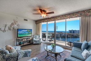 A spacious living room with a large TV, comfortable seating, a balcony with a view of the city, and a ceiling fan. - Terrace At Pelican Beach 0804 3 Bedroom Condo (Destin)