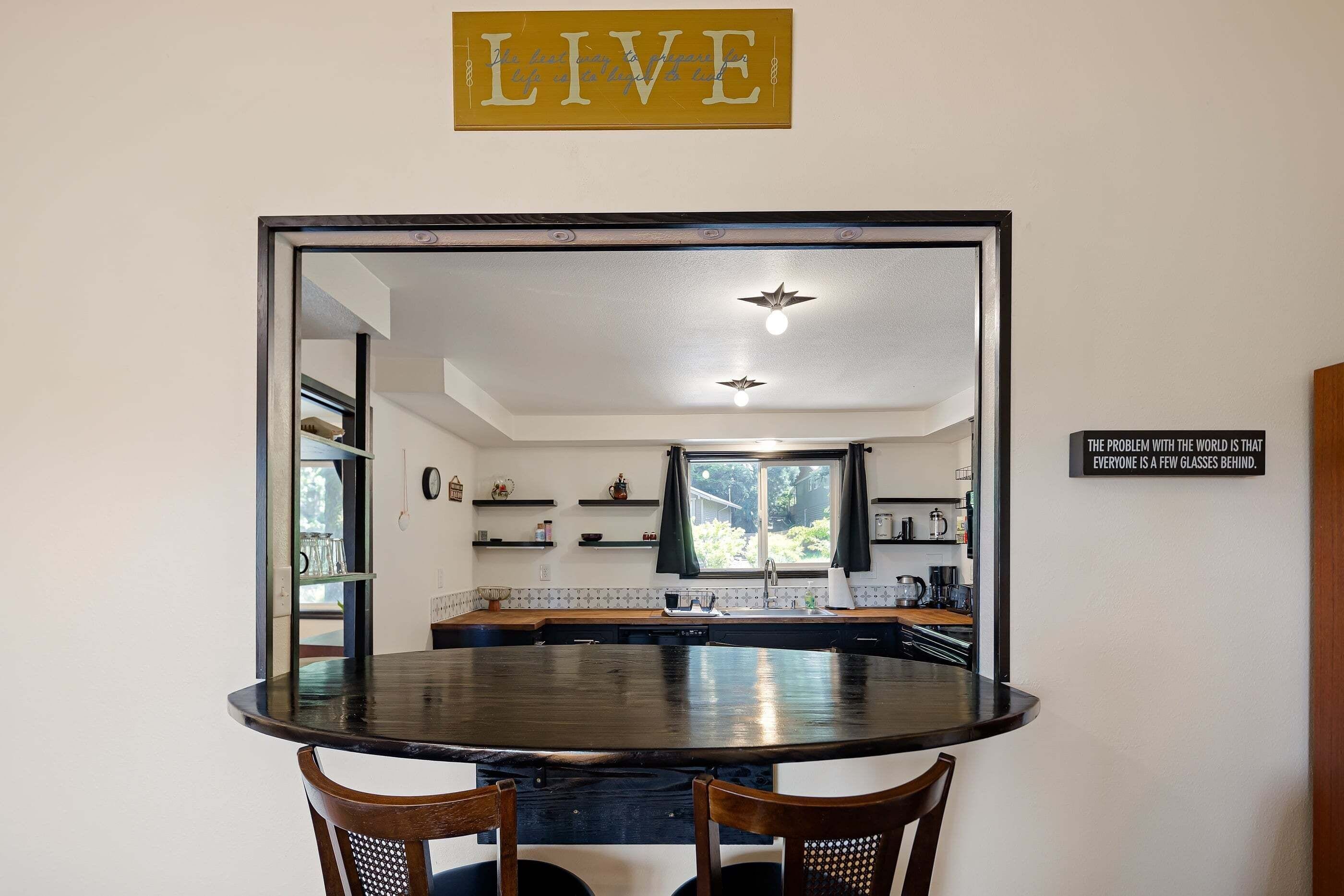 A dining area with a round table and chairs, a kitchen visible through an open door, and a motivational sign on the wall.