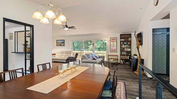 A dining area with a wooden table, chairs, and a chandelier. There is a sofa and a bookshelf in the background.