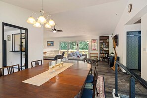 A dining area with a wooden table, chairs, and a chandelier. There is a sofa and a bookshelf in the background. - Wake Robin House - Comfortable & Close To Campus 4 Bedroom Home (Eugene)