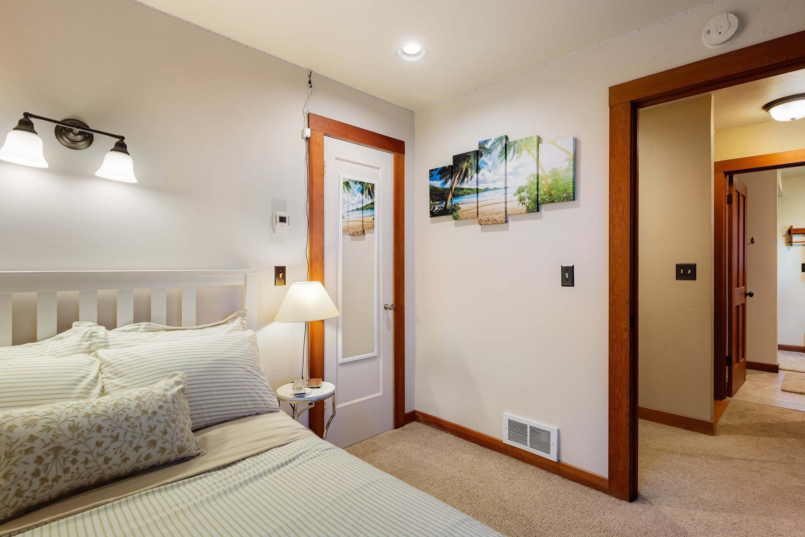A hotel room with a bed, a nightstand, a lamp, a painting, and a door leading to another room.