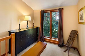 Una habitación de esquina con un banco de madera, un aparador oscuro con dos velas encendidas, una lámpara y una ventana con cortinas.