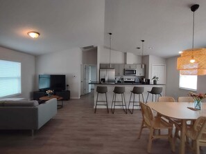 A modern living room with a sofa, TV, and coffee table. A dining area with a table and chairs is adjacent to a kitchen with a microwave and refrigerator. - Moments From The Trail Pool Villa 3 Bedroom Home (Inverness)