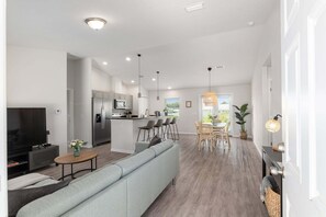 A modern living room with a grey sofa, a TV, and a dining area with a table and chairs. - Moments From The Trail Pool Villa 3 Bedroom Home (Inverness)