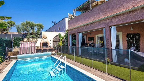 Ein Swimmingpool mit Glasverkleidung, eine überdachte Terrasse und ein Haus mit rosa Fassade.