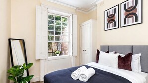 Ein ordentlich bezogenes Bett mit weißer und dunkelblauer Bettwäsche, ein grauer Kopfteil und ein Fenster mit weißen Fensterläden.