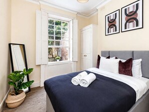 Ein ordentlich bezogenes Bett mit weißer und dunkelblauer Bettwäsche, ein grauer Kopfteil und ein Fenster mit weißen Fensterläden.