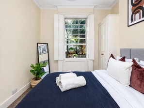 Une chambre à coucher avec un lit, une fenêtre donnant sur un paysage verdoyant, une plante en pot et un tableau encadré accroché au mur.