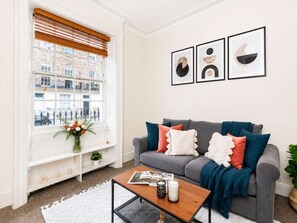 Un salon avec un canapé gris, une table basse et une fenêtre avec des stores. Trois tableaux encadrés sont accrochés au mur.