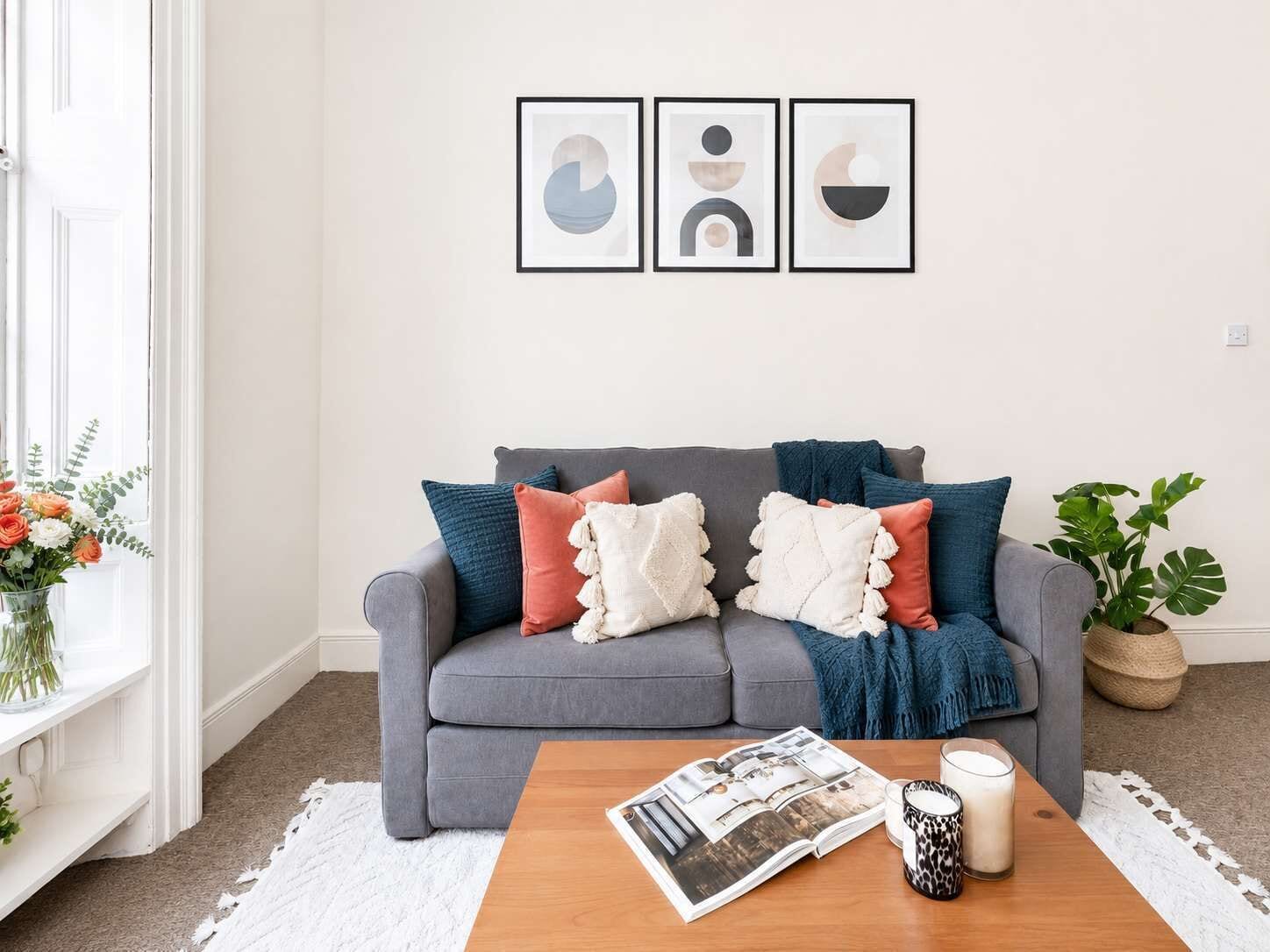 A living room with a grey sofa, colorful cushions, a coffee table with a magazine, a candle, and a plant.