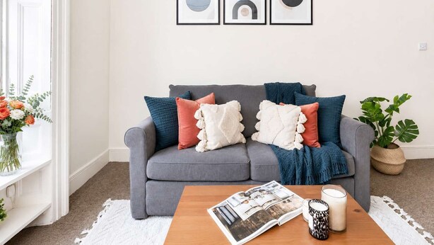 Ein Wohnzimmer mit grauem Sofa, bunten Kissen, einem Couchtisch mit einer Zeitschrift, einer Kerze und einer Pflanze.