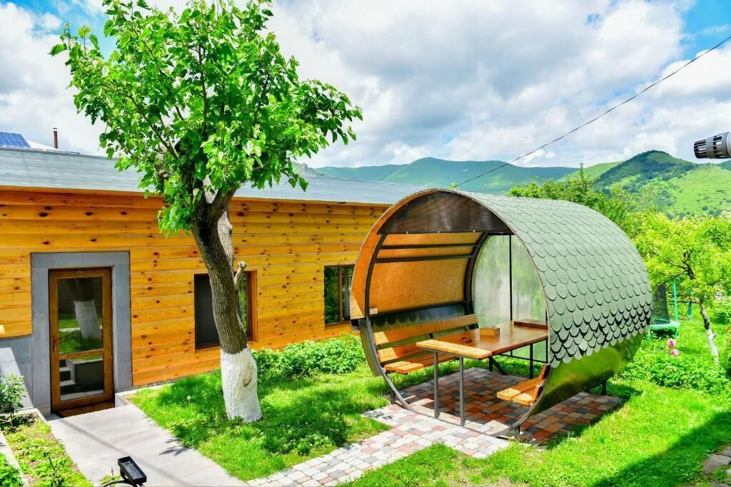 A modern wooden house with a unique barrel-shaped structure attached, a tree in the yard, and a view of mountains in the background.