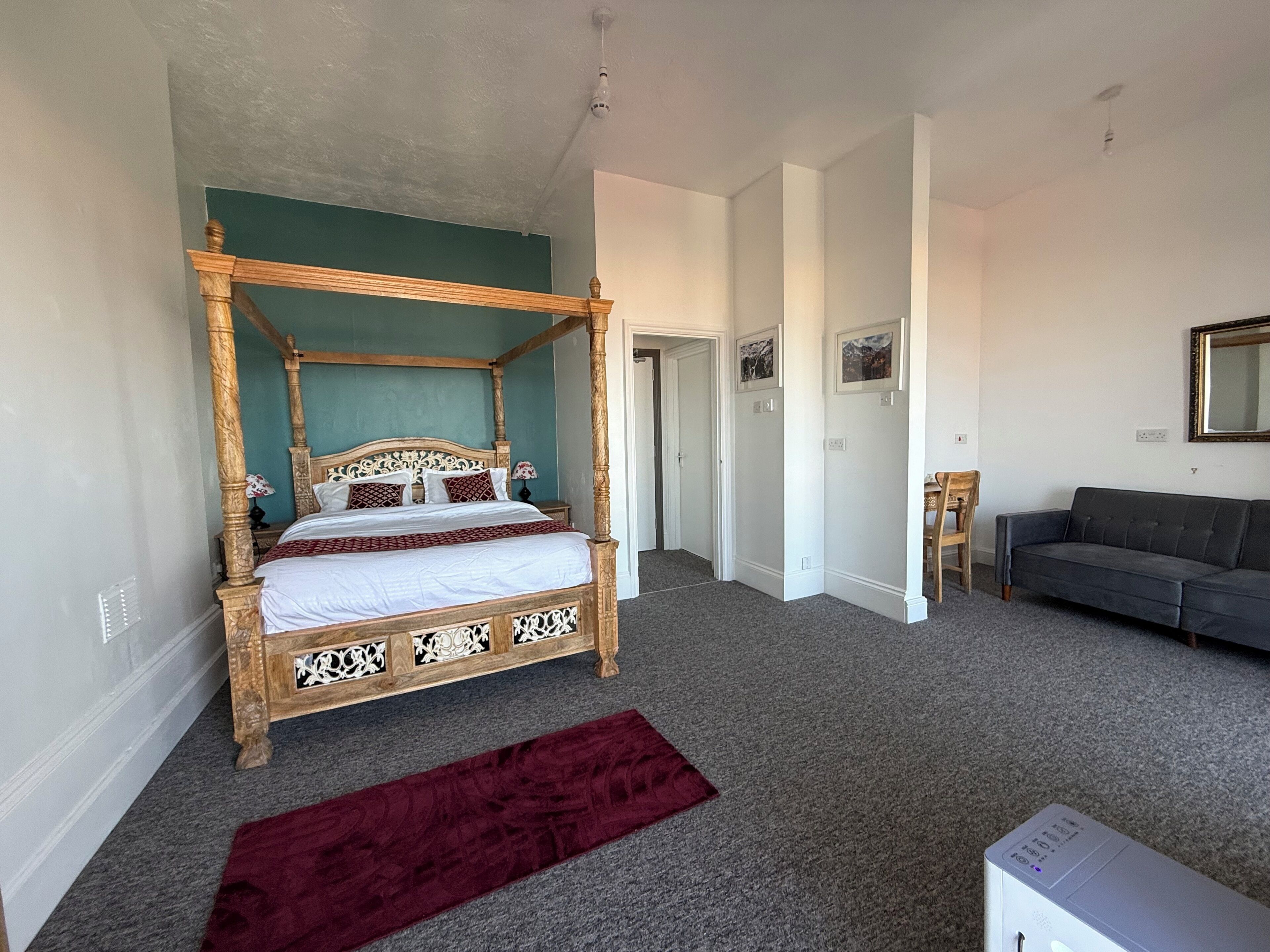 A four-poster bed with a wooden frame and white bedding, a grey sofa, and a red runner on the floor.