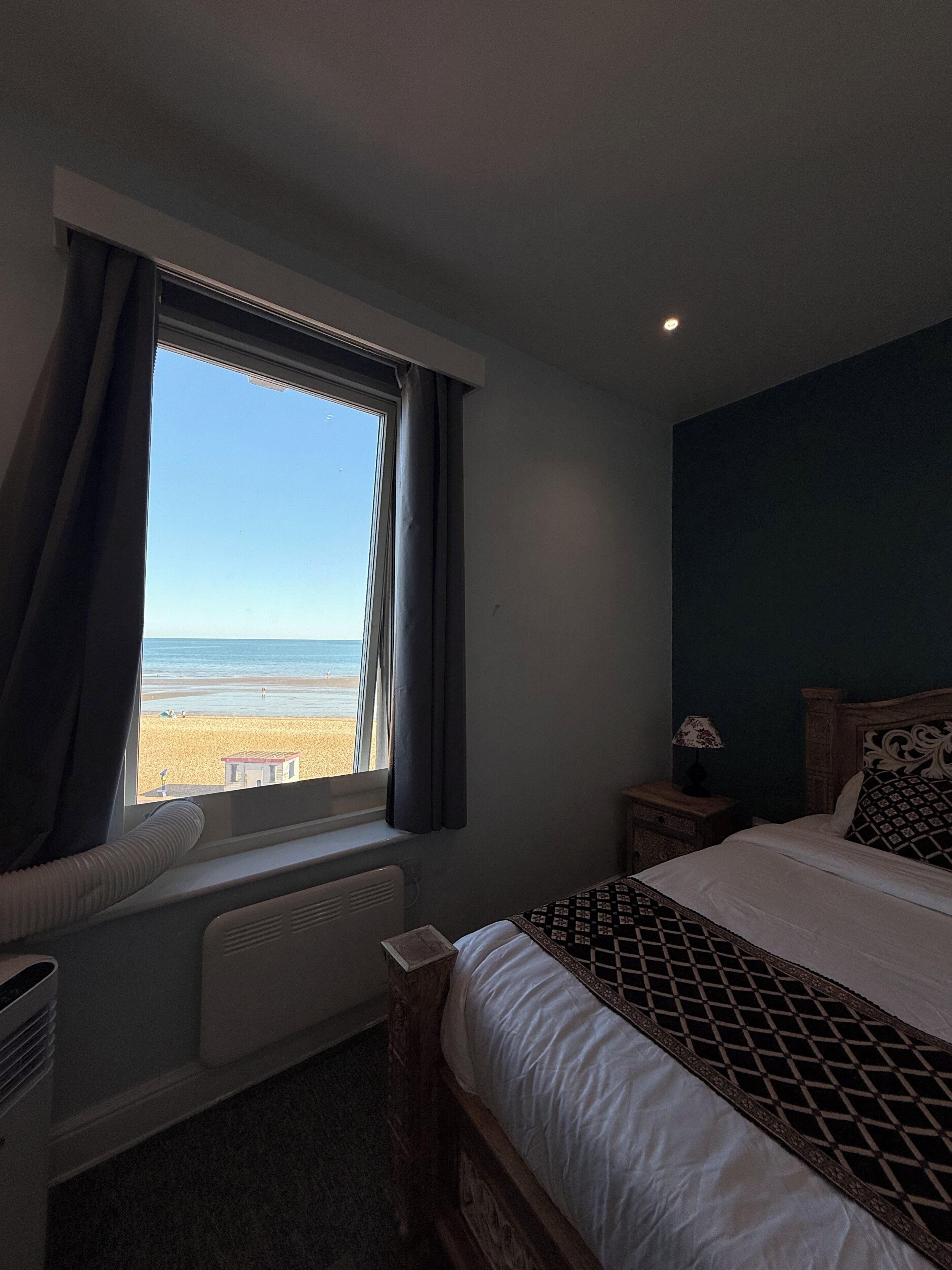A hotel room with a bed, a bedside table with a lamp, a window with curtains, and a view of the beach.