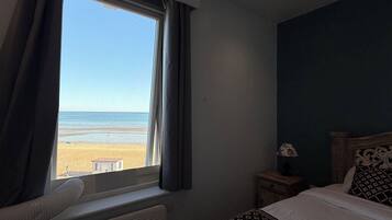 A hotel room with a bed, a bedside table with a lamp, a window with curtains, and a view of the beach.