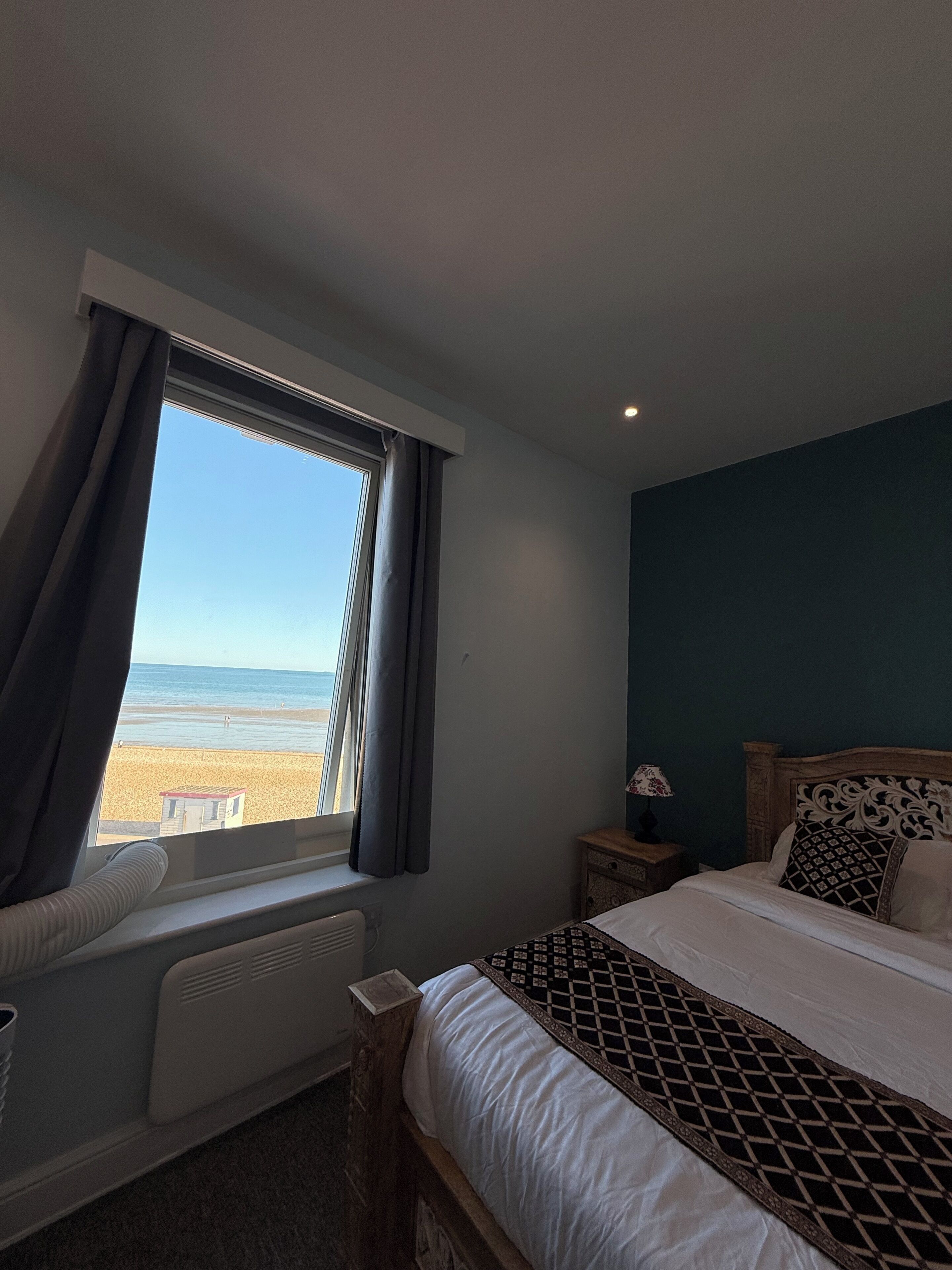 A bedroom with a bed, a nightstand, a window with curtains, and a view of the beach.