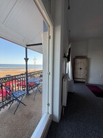 Un umbral abierto que da a un balcón con vistas a la playa, una mesa, sillas y un mueble decorativo.