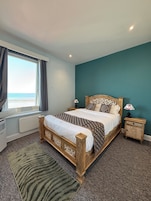 Un dormitorio con cama, mesita de noche, l ámpara, ventana con cortinas y vista a la playa.