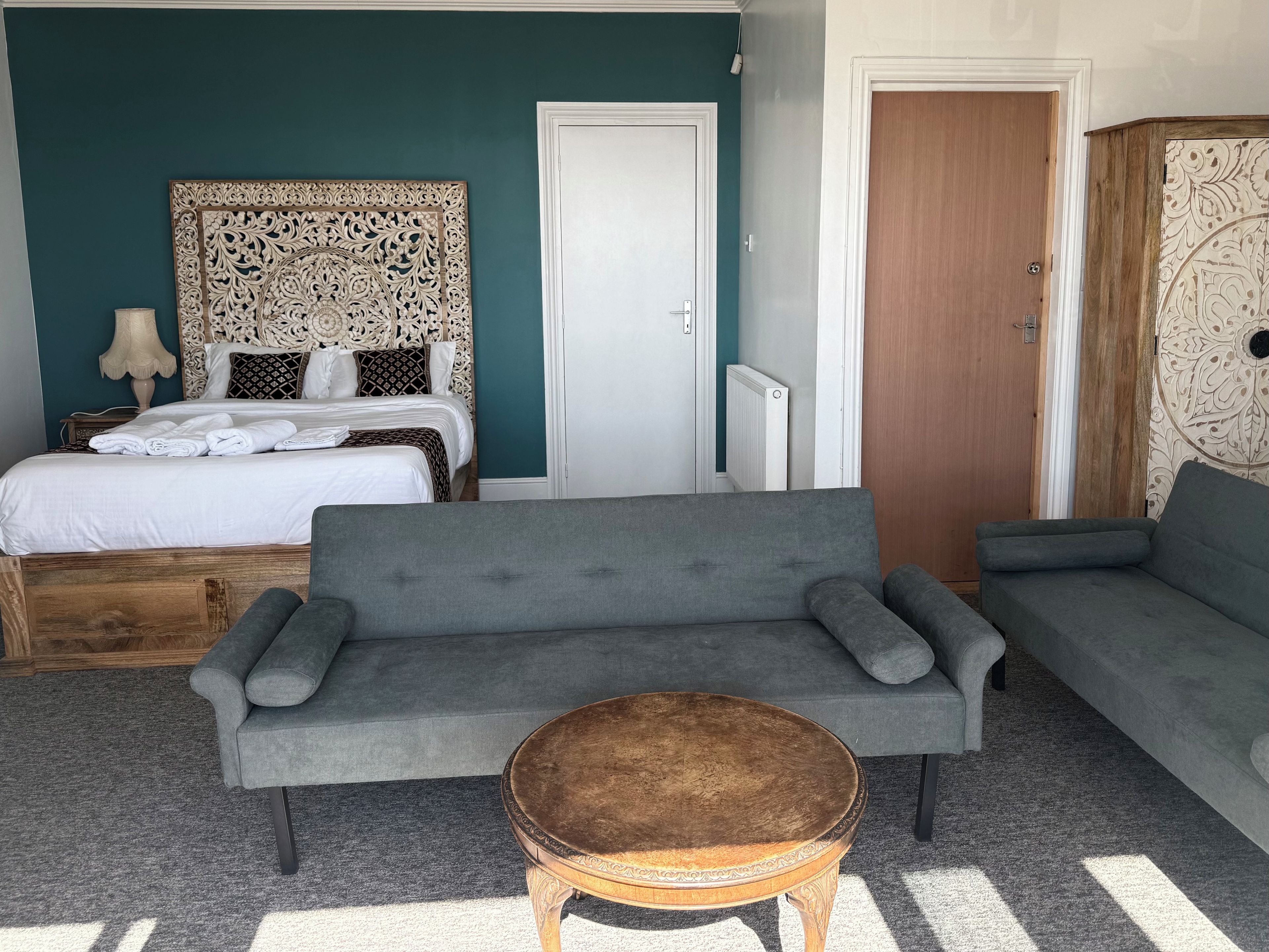 Ein Schlafzimmer mit einem Bett, einem grauen Sofa, einem runden Holztisch und einem Kleiderschrank mit gemusterten Türen.