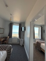 A hotel room with a bed, a desk, a chair, a heater, a TV, and a window with a view of the beach.