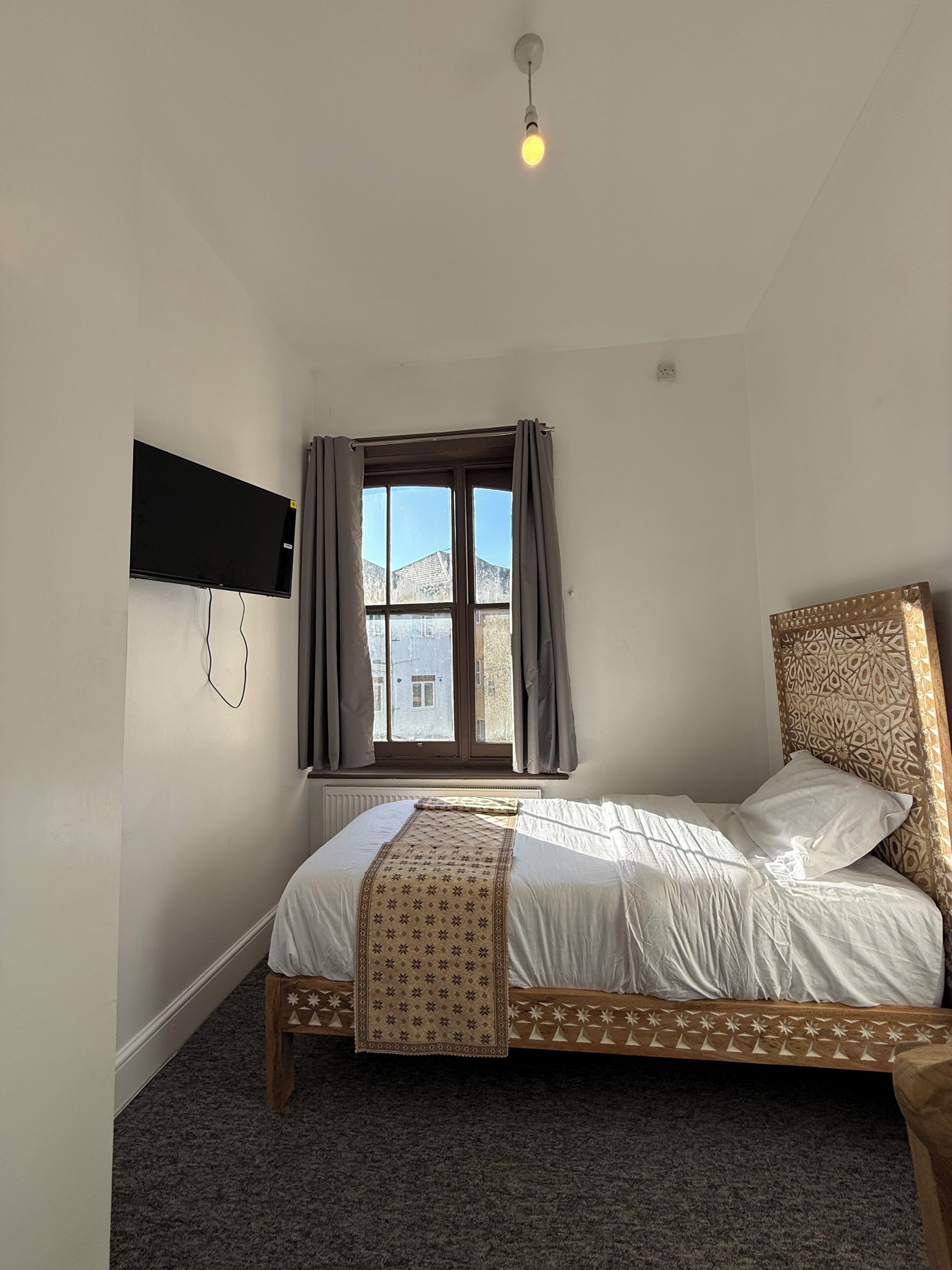 A bedroom with a bed, a window with curtains, a flat-screen TV mounted on the wall, and a light fixture.
