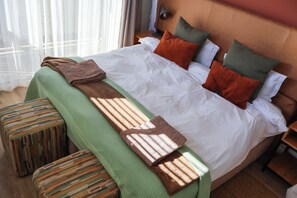 Una cama bien hecha con una manta verde y un plaid marrón, un banco de madera y una mesita de noche con una lámpara.