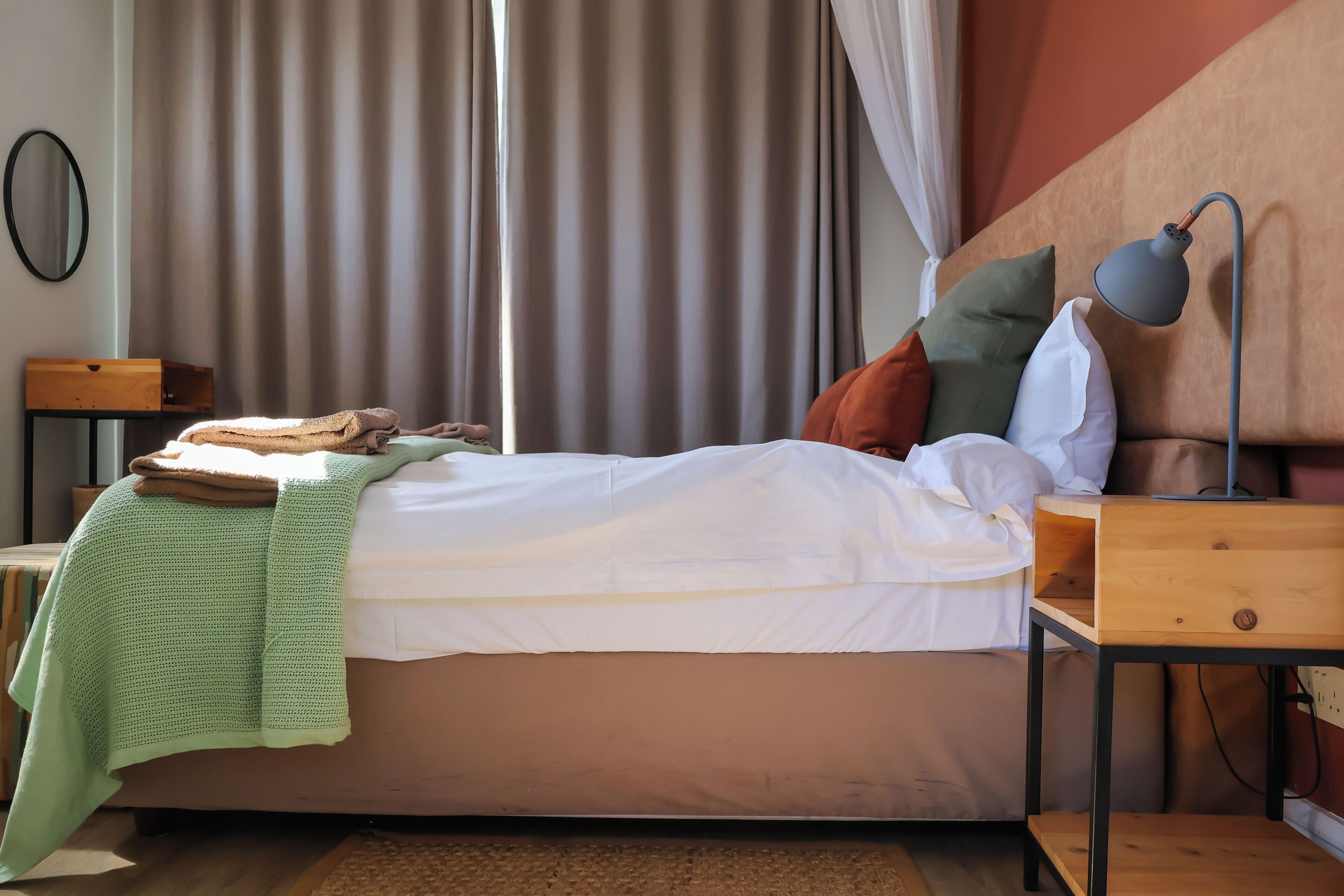 Een netjes opgemaakt bed met een witte deken en kussens, een bedlampje en een houten nachtkastje met een lamp.